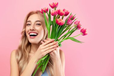 Mutlu Kadın Günleri. Pembe stüdyo arka planında elinde lale buketi tutan kokteyl elbisesi giymiş heyecanlı genç bayan. Bahar tatili için çiçekli neşeli sarışın manken.