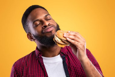 Afrikalı Milenyum Adamı Lezzetli Burger Sarı Stüdyo Arkaplanda Duruyor. Siyah adam hamburger yemekten zevk alıyor. Sağlıksız beslenme alışkanlığı, aşırı yeme. Abur cubur aşığı kavramı