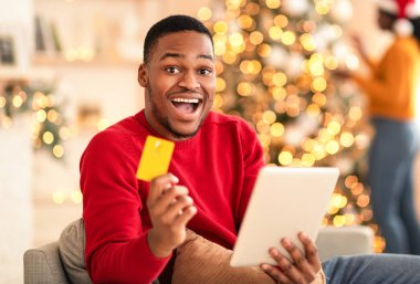Mağazadan, nakitten ve internetten süper bir teklif. Başarılı Afrika kökenli Amerikalı koca, oturma odasında tablet ve kredi kartı taşıyor. Noel Baba şapkalı kadın, Noel Baba ağacı süslemesi yapıyor.