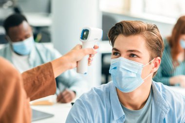 Dijital tıbbi temassız kızılötesi termometre kullanan kadın vücut ısısını ölçüp kontrol ediyor koruyucu maskeli erkek öğrencileri tarıyor bulaşıcı hastalıkların yayılmasını önlüyor.