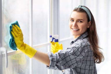 Deterjan ve cam temizleme bezi ile gülümseyen genç kadın. Neşeli milenyum hanımı ev işlerini yapıyor, ev işlerini yapıyor. Profesyonel sağlık hizmeti kavramı