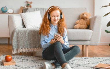 Boş zaman kavramı. Gülümseyen kızıl saçlı liseli kız evde koltuğa yaslanıyor, kablosuz kulaklıkla müzik dinliyor ve cep telefonuyla sohbet ediyor, boş zamanlarının tadını çıkarıyor.