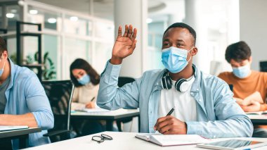 Salgın Konsepti Dönemi Eğitimi. Üniversitedeki konferansta virüs hastalıklarından korunmak için cerrahi maske takan siyah bir öğrencinin portresi cevap ya da soru sormak için elini kaldırıyor.