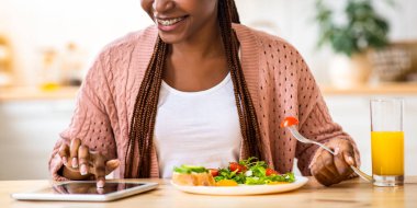 Mutfakta kahvaltı yaparken dijital tablet kullanan gülümseyen siyah kadın, masada oturan, sosyal ağları kontrol eden veya internetten alışveriş yapan Afrikalı Amerikalı olumlu kadın,
