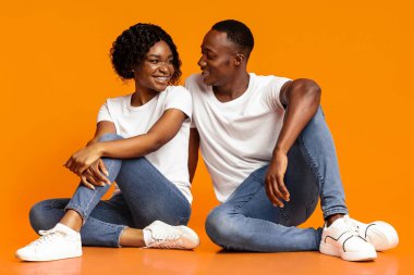 Mutlu Afro-Amerikan aşıklar, turuncu stüdyo arka planında oturup birbirlerine bakıyorlar, kopyalama alanı. Genç siyahi erkek ve kadını sevmek birlikte zaman geçirmek, kaynaşmak.