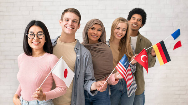 Exchange students and language school for different people. Funny digital young international pupils holding flags of USA, Japan, Turkey, Germany and France in classroom, studio shot, empty space