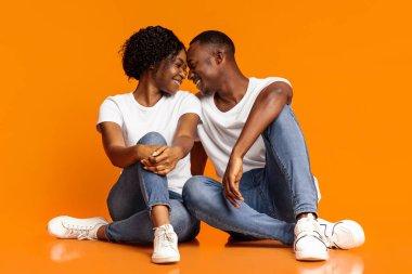 Duygusal siyahi aşıklar genç adam ve kadın turuncu stüdyo arka planında sarılıyorlar, yerde oturuyorlar, alınlarıyla birbirlerine dokunuyorlar ve gülüyorlar, eğleniyorlar, birlikte zaman geçiriyorlar, boşluğu kopyalıyorlar.