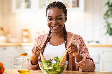 Mutfakta taze sebze salatası hazırlayan mutlu, gülümseyen Afrikalı Amerikalı kadın, masada oturan ve malzemeleri kaseye karıştıran, evde yemek yapmaktan keyif alan, uzay kopyalayan genç, siyah kadın.