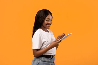Modern dijital tabletli, turuncu stüdyo arka planında poz veren pozitif, genç siyahi kadın, kopyalama alanı. Neşeli Afrikalı kadın cep telefonu uygulamasını ped, teknoloji ve eğlence konseptinde kullanıyor.