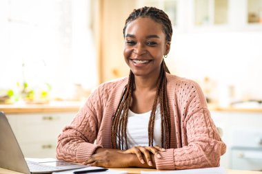 Mesafe işi. Dizüstü bilgisayarla masada oturan mutlu siyahi kadın portresi, masadaki bilgisayarda çalışan neşeli Afrikalı Amerikalı kadın, kameraya gülümseyen, boş alan...