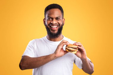 Elinde hamburgerle kameraya gülümseyen mutlu siyah adam sarı arka planda duruyor. Stüdyo çekimi. Abur cubur ve aldatma yemeği, beslenme ve erkek diyeti konsepti. Aşırı yeme alışkanlığı