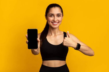 Sağlıklı Yaşam İçin Uygulama. Portakal rengi stüdyo arka planında izole edilmiş, işaret işaretini gösteren, siyah ekranlı cep telefonu tutan gülümseyen sporcu kadının portresi.