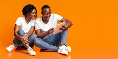 Neşeli siyah çift yerde birlikte oturuyor ve dijital tablet kullanarak turuncu stüdyo arka planında fotokopi çekiyorlar. Afrikalı Amerikalı mutlu kadın ve erkek dijital ekranda oyun oynuyorlar.