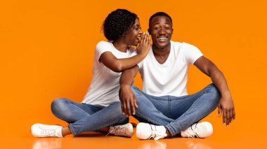 Zenci kadın heyecanlı erkek arkadaşına turuncu stüdyo arka planında sırlar fısıldıyor, boşluk. Afrikalı Amerikalı kadın, meraklı erkeğiyle dedikoduları paylaşıyor, yerde birlikte oturuyor, eğleniyor.