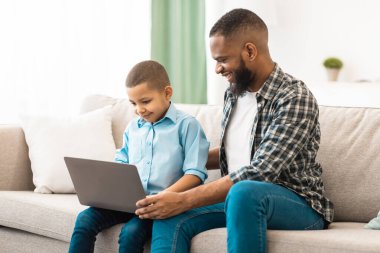 Mutlu siyah baba, oğluna dizüstü bilgisayar kullanmayı öğretiyor koltukta oturuyor ve internette birlikte geziniyor. Teknoloji ve Gadget, Baba Kavramı ile Hafta Sonu