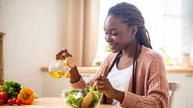 Mutfakta sağlıklı sebze salatası hazırlayan mutlu genç bayan, bovlinge zeytinyağı ve baharat ekliyor, gülümseyen Afro-Amerikan kadın evde yemek pişirmekten hoşlanıyor, bedava uzay.