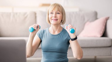 Evde online spor eğitimi. Son sınıf öğrencisi çekici bir kadın oturma odasında dizüstü bilgisayarla egzersiz yapıyor. Kol kasları üzerinde çalışan güçlü olgun bir bayan.