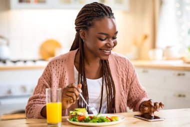 Callories Sayma Uygulaması. Gülümseyen siyahi kadın, evde kahvaltı ederken akıllı telefonuyla modern uygulamaları kullanıyor, masada oturan mutlu Afrikalı kadın, günlük beslenme verilerini kontrol ediyor.