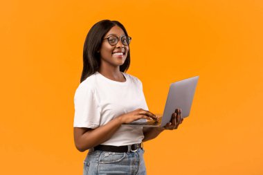Gözlüklü, yeni dizüstü bilgisayarı olan, gülümseyen, turuncu stüdyo geçmişi olan, fotokopi alanı olan bir panorama olan mutlu Afrikalı Amerikalı genç kadın. Neşeli siyahi kız öğrenci defter kullanıyor, internetten çalışıyor.