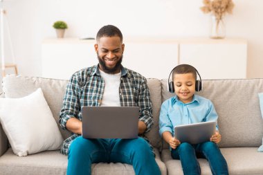 Black Father ve Little Boy bilgisayarlar kullanarak dizüstü bilgisayarları ve dijital tabletleri tarıyor evdeki koltukta oturan kablosuz kulaklıkları takıyor. Aile Parçacıkları Konsepti