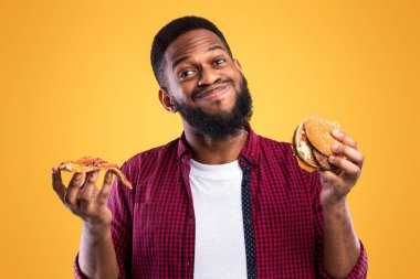 Gülümseyen Afrikalı Adam Burger ve Pizza Pozisyonu Sarı Arkaplanda Stüdyoda Duruyor. Abur cubur yiyenler portresi. Sağlıksız Fast food ve Erkek Beslenme, Aldatma ve Aşırı Yeme