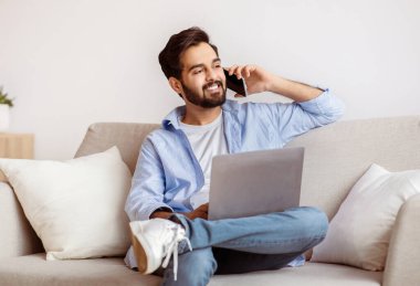 Uzaktan Çalışma Konsepti. Yakışıklı Arap erkek, dizüstü bilgisayar kullanıyor ve cep telefonuyla konuşuyor oturma odasında bilgisayarla oturuyor, uzak iş fırsatlarının tadını çıkarıyor, boşluğu kopyalıyor.