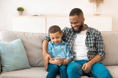 Siyah çocuk ve babası cep telefonu kullanıyor internette oyunlar oynuyor koltukta oturuyor ve oturma odasında interneti inceliyor. Smartphone, Family and Gadgets Concept için yeni uygulama