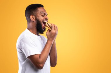 Siyah Millennial Guy Burger 'ı Isıran Adam Sarı Stüdyo Arkaplanı Üzerinde Hurda Yiyen Adam. Yan Görünüm, Boşluk Boşluğu. Afro-Amerikan adam lezzetli çizburgerin tadını çıkarıyor. Aşırı yeme, Sağlıksız Beslenme alışkanlığı