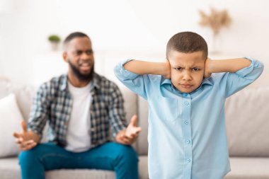 Kızgın baba evde dikilip kameraya bakarken siyah çocuk kulaklarını tıkıyor. Aile içi şiddet, mutsuz çocukluk, aile sorunu ve kötü ilişki. Seçici Odaklanma