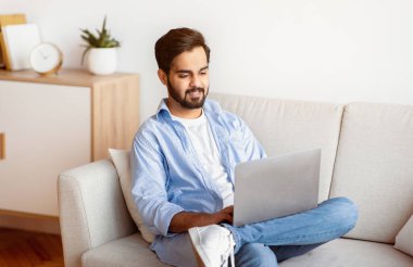Doğu Milenyum Erkeği evde koltukta dinlenirken dizüstü bilgisayar kullanıyor. Yakışıklı Arap Serbest Çalışan Adam bilgisayarla koltukta oturuyor, uzaktan çalışıyor ya da internetten video izliyor, Boşluğu Kopyala