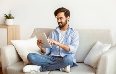Doğu Milenyum Adamı dijital tabletle evde vakit geçiriyor, genç Arap adam oturma odasında oturuyor, internetten alışveriş yapıyor, internete bakıyor ya da uzaktan çalışıyor, ev ile eğleniyor.