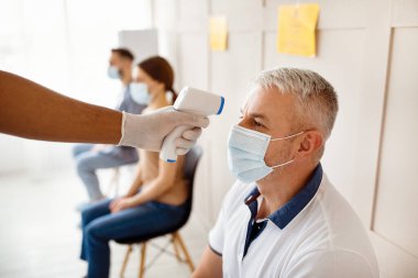Hastanede kovid 19 aşısı bekleyen tıbbi maskeli kıdemli bir adam. Doktor ateşini kızılötesi termometreyle ölçüyor. Coronavirus 'a karşı bağışıklık, nüfus koruması