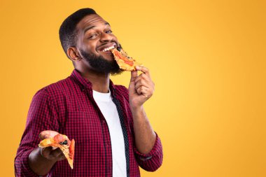 Sarı Stüdyo Arkaplanının Üzerinde Duran Lezzetli Pizza Yiyen Zenci Adam. Abur cubur fazla yeme. Fast food yiyici, sağlıksız pizzayı ısırıyor. Beslenme ve yeme alışkanlığı kavramı