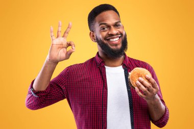 Neşeli Afro-Amerikalı Adam Burger 'lı Poz Veriyor Tamam tabelası Sarı Stüdyo Arkaplanda Duran Kameraya Gülümsüyor. Siyah Adam Hurda Yemeği Onaylıyor