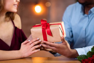 Sevgililer Günü Kutlaması. Loving Black Man 'in restoranda romantik bir akşam yemeğinde mutlu kız arkadaşına hediye paketi vermesi, yıldönümü kutlamaları, güzel hediyeler, kırpılmış görüntüler, yakın çekim...