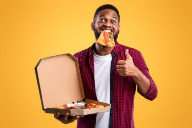 Ağzında pizza dilimi olan mutlu siyah adam sarı stüdyo arka planında duran pizzacıya gülümseyen başparmağını kaldırıyor. Harika Abur cubur ve Hile Yemekleri
