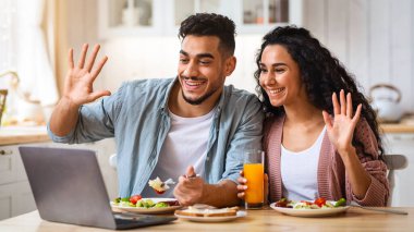 Neşeli Arap çift, birlikte kahvaltı ederken, mutfakta dizüstü bilgisayarla video görüşmesi yapıyor, kameraya el sallıyor, arkadaşlarıyla sohbet ediyor. Ortadoğu eşleri çevrimiçi iletişimden keyif alıyorlar