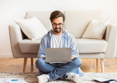 Uzaktan Çalışma Konsepti. Gülümseyen Arap Serbest Çalışan Adam evde dizüstü bilgisayar kullanıyor, oturma odasında yerde oturuyor, Doğulu genç adam uzaktan ve uzaktan çalışma fırsatlarını değerlendiriyor,