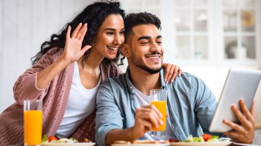 Modern Müslüman çift mutfakta dijital tablet üzerinde video görüşmesi yapıyor, birlikte kahvaltı ederken, gülümseyen orta doğulu çift kameraya el sallıyor, çevrimiçi iletişimin tadını çıkarıyorlar, bedava alan.