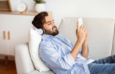 - Boş zaman. Akıllı telefonlu yakışıklı, kablosuz kulaklıklı, oturma odasında dinlenirken müzik dinleyen, gülümseyen Arap adam, evde dinlenen, ses yayınının keyfini çıkaran...