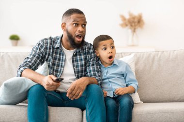 Televizyon programlama sorunu. Şok olmuş Afrikalı baba ve oğul TV 'de tartışmalı içeriği izliyorlar evde ağzı açık bir şekilde koltukta oturuyorlar. Ebeveyn Kontrolü Konsepti