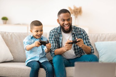 Neşeli siyah baba ve oğlu hafta sonu evde koltukta oturup video oyunu oynuyorlar. Genç Afrikalı baba çocuğuyla vakit geçirmek için oyunlar oynuyor.