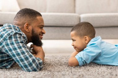 Mutlu Afrikalı baba ve oğul evde yerde yatarken birbirlerine bakıyorlar. Baba ve Çocuk Kaynaşması. Çocuk ve Babalar İlişki ve Güven Konsepti. Yan Görünüm Portresi