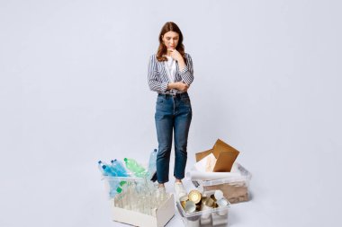 Etrafında çeşitli atık konteynırlara bakan dalgın genç bir kadın, geri dönüşüm için farklı malzemelerle kutular arasında duran düşünceli bir kadın, ışık stüdyosunun arka planında poz veren, boşluğu kopyalayan...