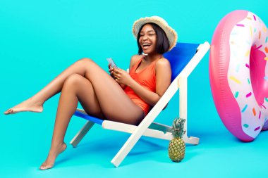 Güzel siyah kadın şezlongda dinleniyor, akıllı telefon kullanıyor, kumsalda arkadaşlarıyla sohbet ediyor, mavi arka planda. Yaz tatili boyunca internette iletişim kuran neşeli genç bayan.