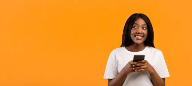 Modern akıllı telefon kullanarak ve fotokopi alanına bakarak, heyecan verici reklamlar okuyarak, turuncu stüdyo arka planıyla, panoramayla mutlu, siyah bir bayan. Mobil uygulama kavramı