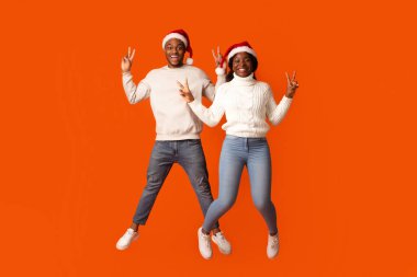 Noel Baba Şapkalı Afro-Amerikan eşleri zıplayarak ve barış jesti göstererek, turuncu stüdyoda birlikte eğlenerek, siyahi çiftler Noel tatillerini kutlarken,