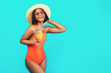 Hasır şapkalı ve mayo giymiş siyah kadın yaz tatilinde tropik meyve kokteyli ile poz veriyor, mavi stüdyo arka planı. Bikinili gülümseyen genç kadın taze içki içiyor, plaj partisinin tadını çıkarıyor.