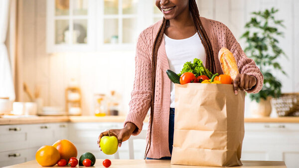 Black Smiling Lady Unpacking Paper Bag With Fresh Organic Vegetables And Fruits After Grocery Shopping, Happy African American Woman Standing In Cozy Kitchen Interior At Home, Copy Space