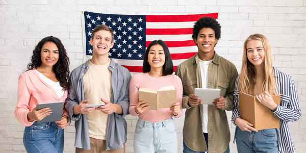 Digital gadgets and books for remote learning English. Happy zoomers multiracial people in casual, read and study, training for test, on white wall background with large flag of America, empty space